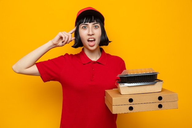 Jovem entregadora caucasiana surpresa segurando recipientes de comida em caixas de pizza