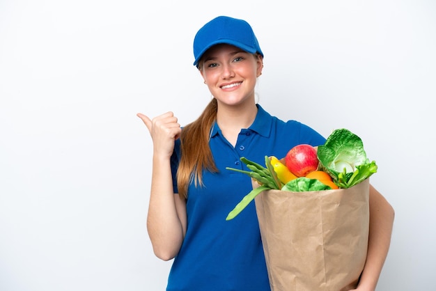 Jovem entregadora levando um saco de comida para viagem isolada no fundo branco, apontando para o lado para apresentar um produto