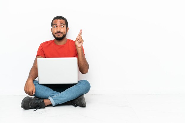 Jovem equatoriano com um laptop sentado no chão isolado em fundo branco com dedos cruzando e desejando o melhor