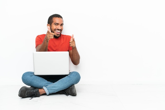 Jovem equatoriano com um laptop sentado no chão, isolado no fundo branco, apontando para a frente e sorrindo