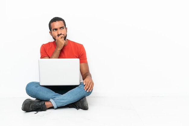 Jovem equatoriano com um laptop sentado no chão isolado no fundo branco com dúvidas e com expressão facial confusa