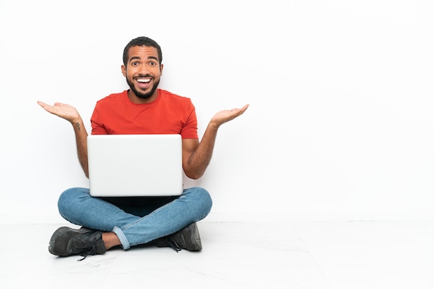 Jovem equatoriano com um laptop sentado no chão isolado no fundo branco com expressão facial chocada