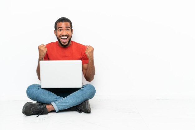 Jovem equatoriano com um laptop sentado no chão isolado no fundo branco comemorando uma vitória na posição de vencedor