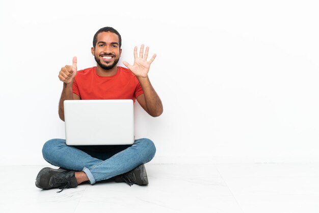 Jovem equatoriano com um laptop sentado no chão isolado no fundo branco contando seis com os dedos