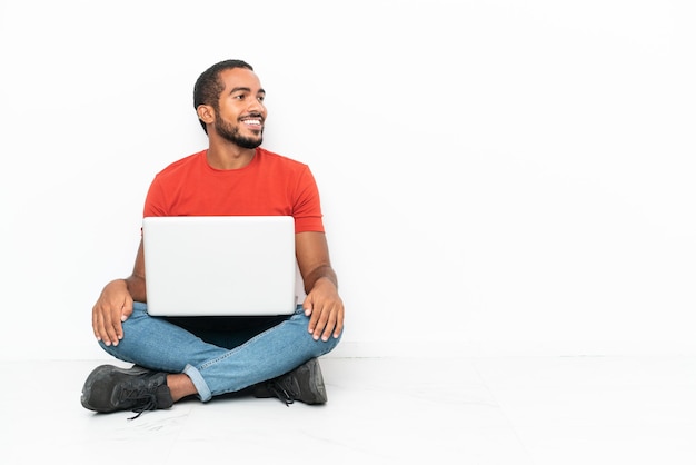 Jovem equatoriano com um laptop sentado no chão, isolado no fundo branco, olhando para o lado