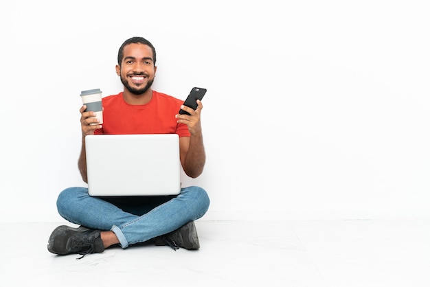 Jovem equatoriano com um laptop sentado no chão, isolado no fundo branco, segurando um café para levar e um celular