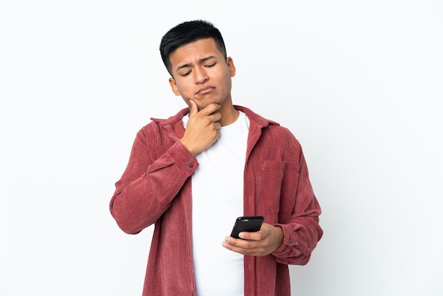 Jovem equatoriano isolado em uma parede branca usando telefone celular e pensando