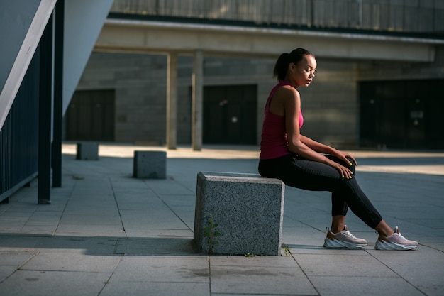 Foto jovem esbelta com roupas esportivas sentada no cubo de pedra na rua e relaxando após o treino