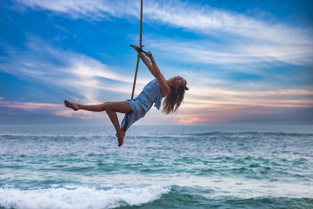 Jovem esbelta de vestido azul balançando no bungee voando sobre o pôr do sol do oceano