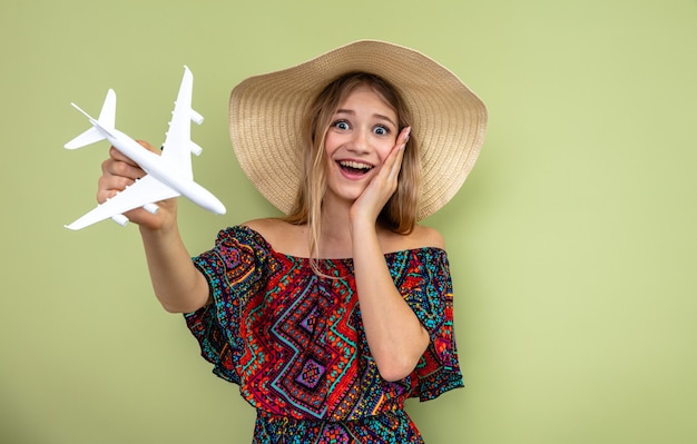 Foto jovem eslava loira surpresa com chapéu de sol, colocando a mão no rosto e segurando o modelo do avião