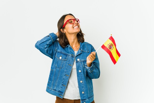 Jovem espanhola segurando uma bandeira isolada na parede branca, sentindo-se confiante, com as mãos atrás da cabeça