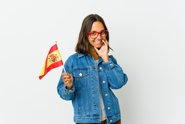 Jovem espanhola segurando uma bandeira isolada no fundo branco, sorrindo feliz e confiante, tocando o queixo com a mão.