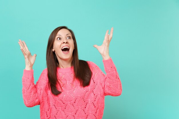 Jovem espantada com uma camisola de malha rosa, mantendo a boca aberta, espalhando as mãos olhando para cima isoladas no fundo da parede azul no estúdio. Emoções sinceras de pessoas, conceito de estilo de vida. Simule o espaço da cópia.