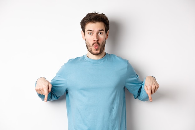 Jovem espantado com a barba, em pé com uma camisa azul casual, dizendo uau e apontando para baixo, olha aqui o gesto, em pé sobre um fundo branco.