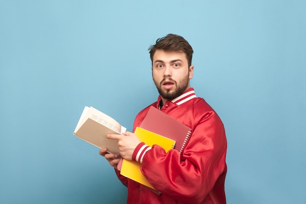 Jovem espantado com barba e livros nas mãos, vestindo uma jaqueta vermelha