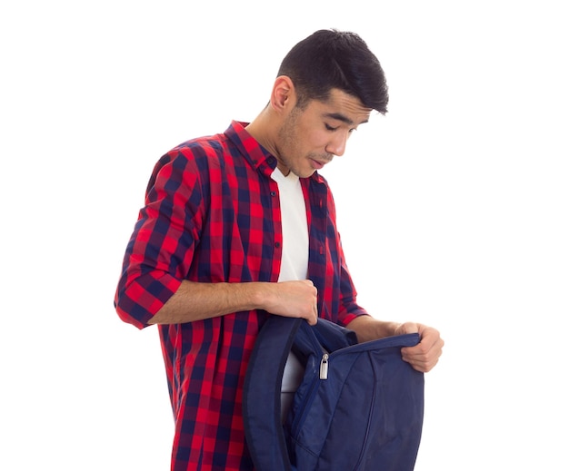 Jovem espantado com cabelo preto em camiseta branca e camisa xadrez vermelha segurando mochila azul