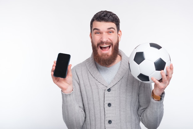 Foto jovem espantado segurando a bola de smartphone e futebol sobre fundo branco