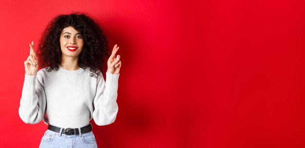 Foto jovem esperançosa com lábios vermelhos e dedos cruzados de cabelo encaracolado para dar boa sorte e fazer desejos rezando