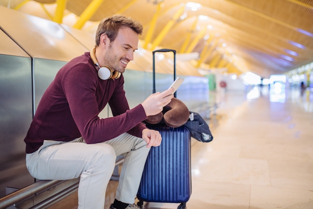 Jovem esperando ouvir música e usando telefone celular no aeroporto com uma mala.