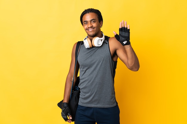 Jovem esporte afro-americano com tranças e bolsa isolada em fundo amarelo saudando com a mão com expressão feliz