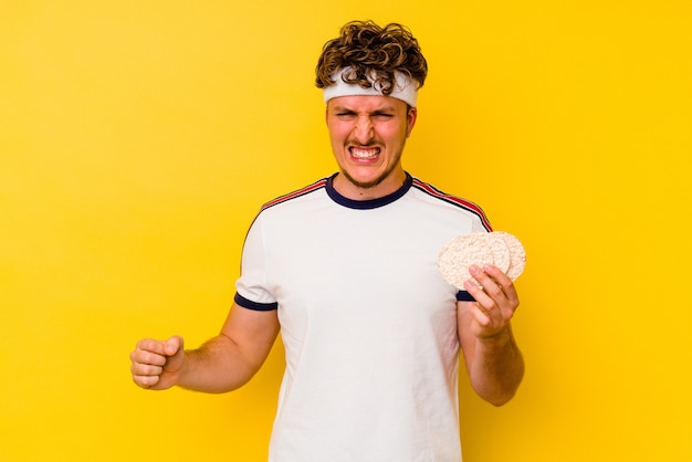 Jovem esporte homem caucasiano comendo um bolo de arroz isolado no fundo amarelo, gritando com muita raiva e agressividade.