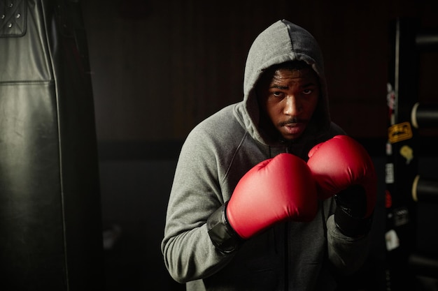 Jovem esportista afro-americano forte em luvas de boxe vermelhas e capuz cinza olhando para a câmera ou saco de pancadas e se preparando para o chute