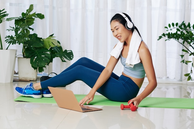 Jovem esportista chinesa sorridente seguindo o tutorial on-line no laptop enquanto se exercita em casa