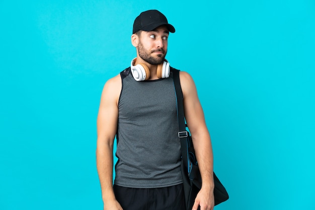 Jovem esportista com bolsa esporte isolada em um fundo azul fazendo gesto de dúvida olhando para o lado