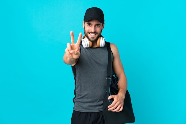 Jovem esportista com bolsa esportiva isolada na parede azul, sorrindo e mostrando sinal de vitória