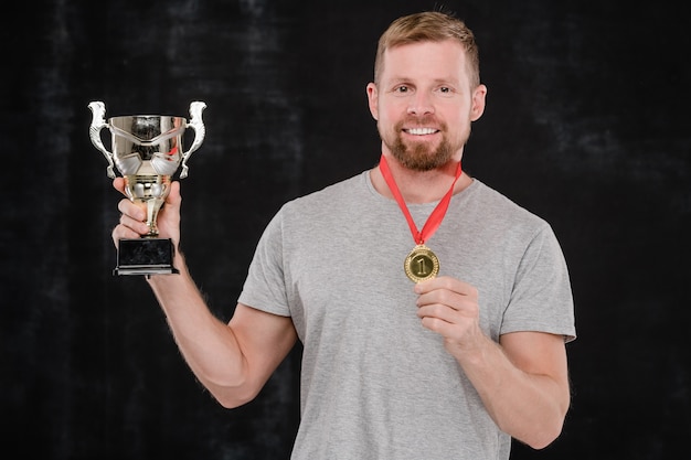 Foto jovem esportista de sucesso mostrando a taça de prata e a medalha de ouro em frente à câmera contra um fundo preto