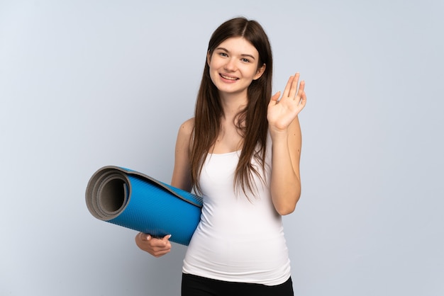 Jovem esportista indo para as aulas de ioga, segurando um tapete e saudando com a mão com uma expressão feliz