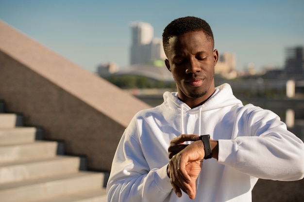 Jovem esportista sério de etnia africana olhando para smartwatch no pulso esquerdo enquanto vai correr pela manhã em ambiente urbano