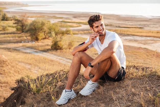 Jovem esportista sorridente, descansando e ouvindo música com fones de ouvido após o treino ao ar livre