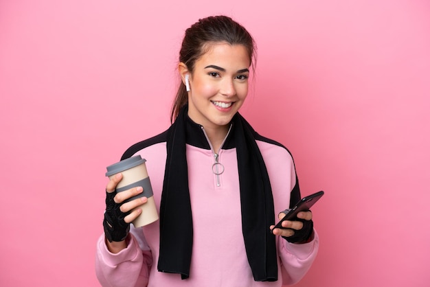 Jovem esportiva brasileira isolada em fundo rosa segurando café para levar e um celular