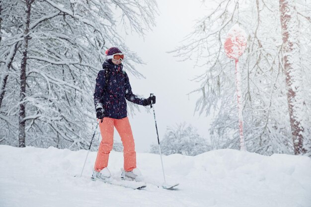 Jovem esquiadora em pé na pista de esqui sob queda de neve segurando bastões de esqui e sorrindo