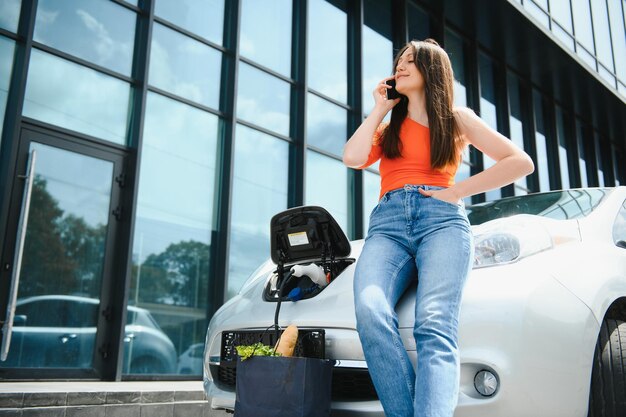 Jovem está de pé perto do carro elétrico e olha para o telefone inteligente O carro alugado está carregando na estação de carregamento para veículos elétricos Compartilhamento de carro