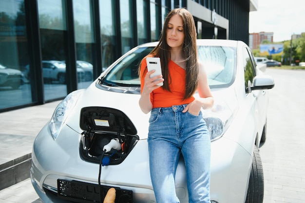 Jovem está de pé perto do carro elétrico O carro alugado está carregando na estação de carregamento para veículos elétricos Compartilhamento de carro