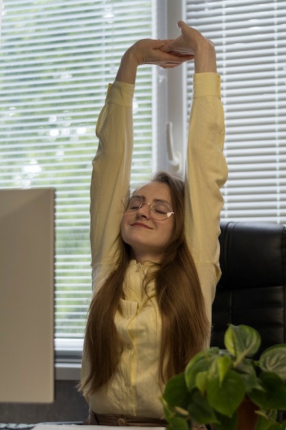 Foto jovem está se alongando no local de trabalho. a menina se senta na cadeira e levanta os braços para esticar as costas. pausa