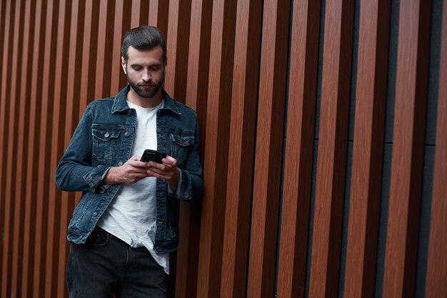 Jovem estilo livre em pé encostado em uma parede de madeira, isolado navegando na internet em smartphone