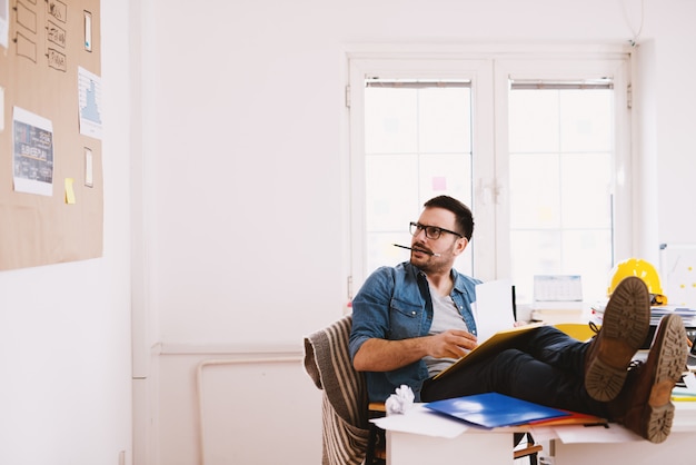 Jovem estressado empresário de homem de negócios moderno com as pernas sobre a mesa do escritório, olhando para o painel de parede com um lápis na boca, enquanto tenta a resolução de problemas.