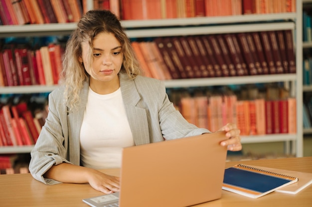 Jovem estudante abre laptop e começa a estudar na biblioteca pública Mulher se preparando para exame universitário