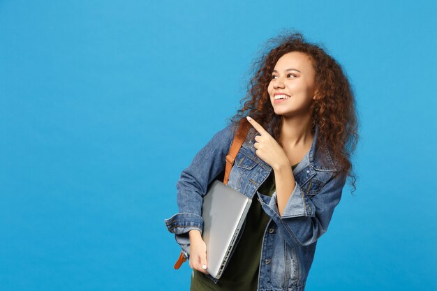 Jovem estudante adolescente afro-americana com roupas jeans, mochila segurando pc isolado na parede azul