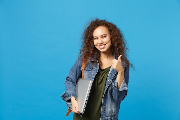 Jovem estudante adolescente afro-americana com roupas jeans, mochila segurando pc isolado na parede azul