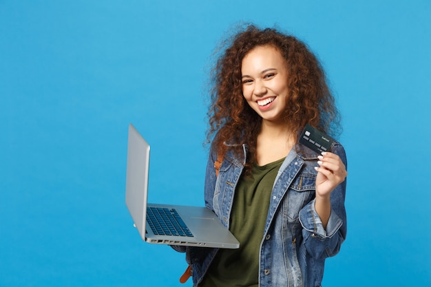 Foto jovem estudante adolescente afro-americana com roupas jeans, mochila trabalhando no pc, segurando o cartão do banco isolado na parede azul