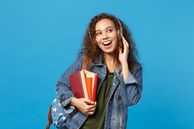 Jovem estudante adolescente afro-americana em roupas jeans, fones de ouvido de mochila isolados na parede azul