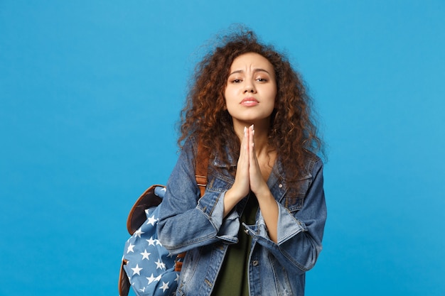 Foto jovem estudante adolescente afro-americana em roupas jeans, mochila fazendo desejo isolada na parede azul