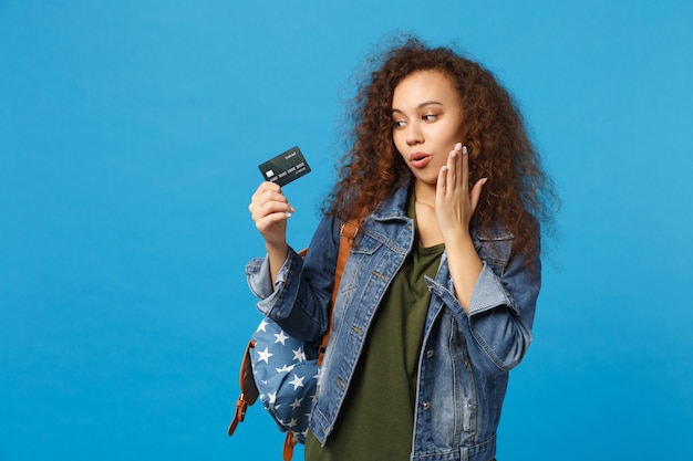 Jovem estudante adolescente afro-americana em roupas jeans, mochila segurando cartão de crédito isolado na parede azul