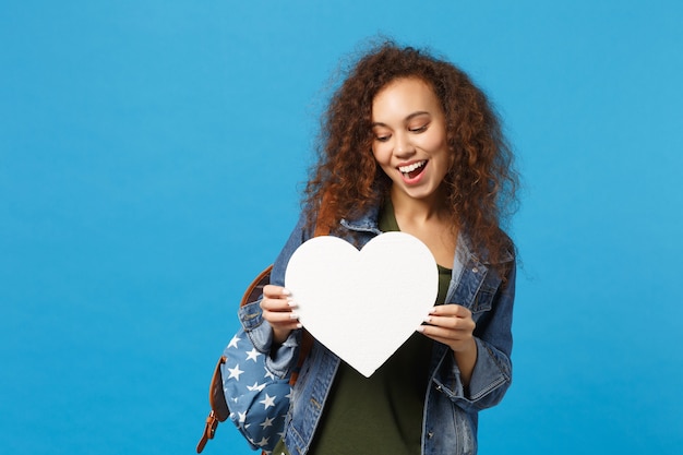 Foto jovem estudante adolescente afro-americana em roupas jeans, mochila segurando coração isolado na parede azul