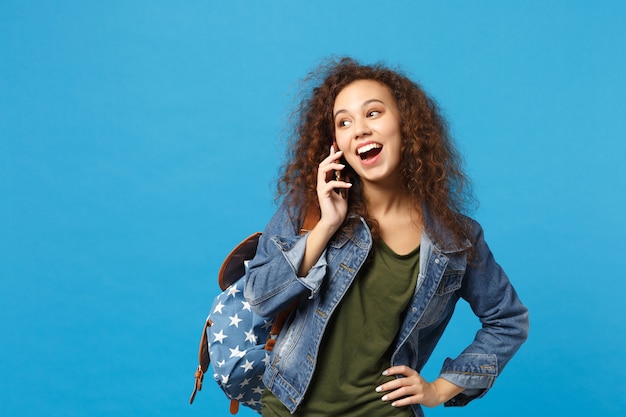 Jovem estudante adolescente afro-americana em roupas jeans, mochila segurando telefone isolado na parede azul