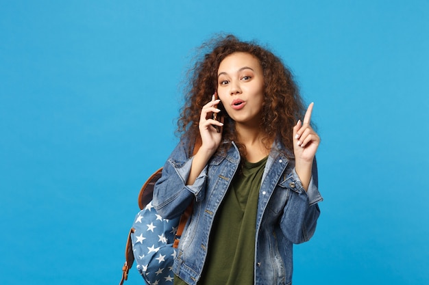 Jovem estudante adolescente afro-americana em roupas jeans, mochila segurando telefone isolado na parede azul